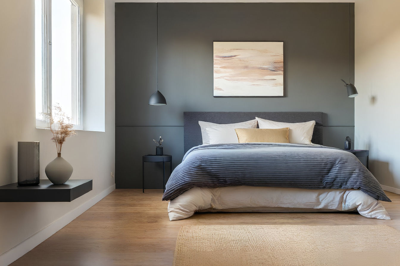 A stylish bedroom features a dark charcoal gray accent wall behind a bed with layered neutral-toned bedding and a textured throw. Two modern bedside lamps with warm light frame the bed, and a piece of abstract art hangs above the headboard. The adjacent walls are painted in a soft white, and the room is filled with natural light from a nearby window. The overall look is sophisticated, cozy, and inviting, highlighting the dramatic impact of the gray accent wall.