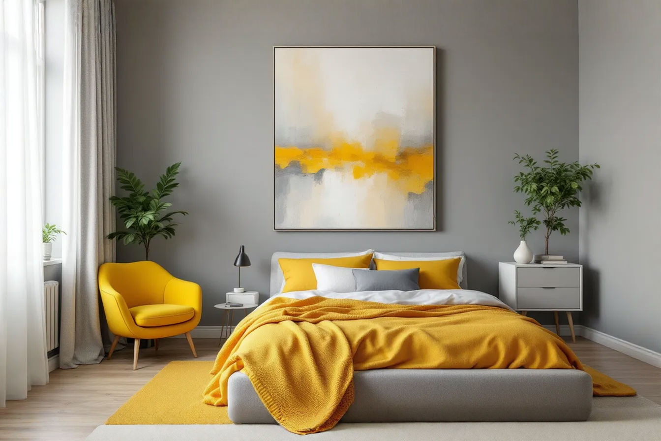 A professionally styled bedroom featuring a large gray upholstered bed with yellow throw pillows and a yellow knitted blanket. The walls are painted in a soft, warm gray. A mustard yellow accent chair sits in one corner. A large abstract painting above the bed incorporates shades of gray, yellow, and white. Natural light streams in from a window with sheer white curtains. The overall mood is balanced, inviting, and modern.