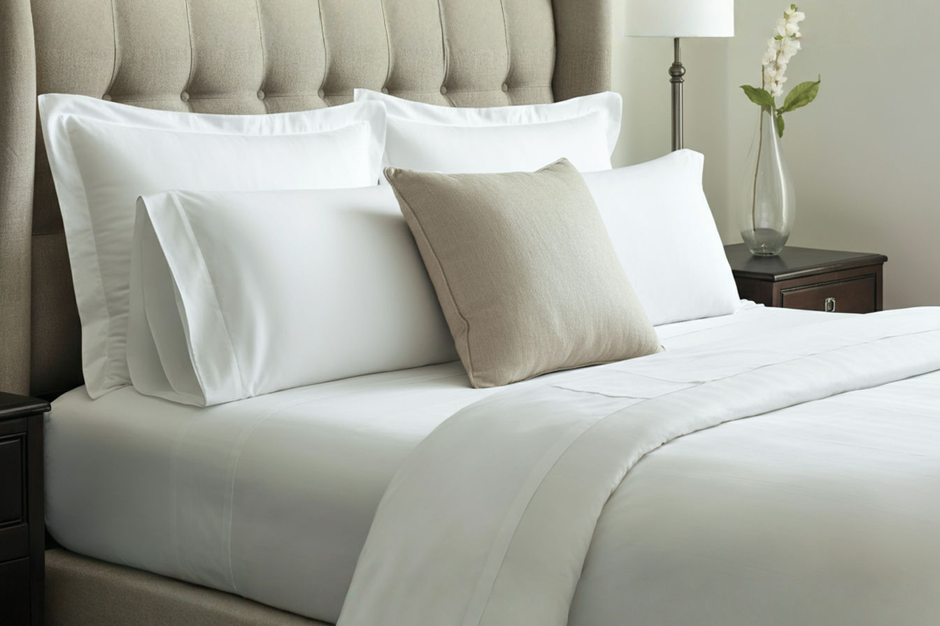 A high-angle professional photograph of a queen-sized bed in a bright, airy bedroom. The bed is neatly made with crisp white sheets and a neutral-colored duvet. The pillows are arranged using the hotel method, showing a layered stack of two sleeping pillows, two Euro shams, and a decorative accent pillow