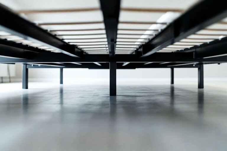 Professional, well-lit photograph showing a close-up view of the underside of a modern, sturdy metal king size bed frame. The image focuses on the central support system, clearly showing thick metal beams and multiple support legs extending to the floor. The perspective is slightly low, emphasizing the strength and density of the support structure. The background is a clean, neutral bedroom floor.